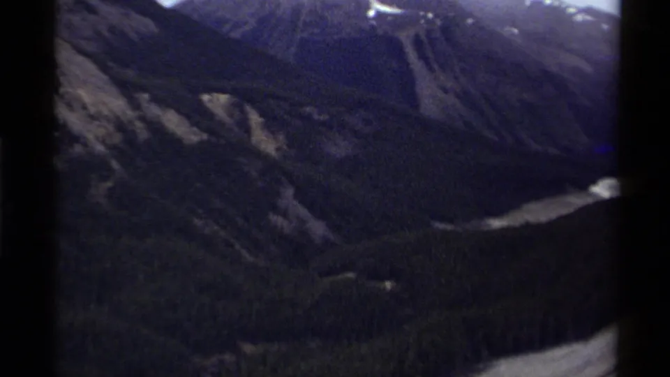 1979 Canada 8mm frame showing a shadowed mountain valley, snow-streaked ridges, forest, and pale water below.