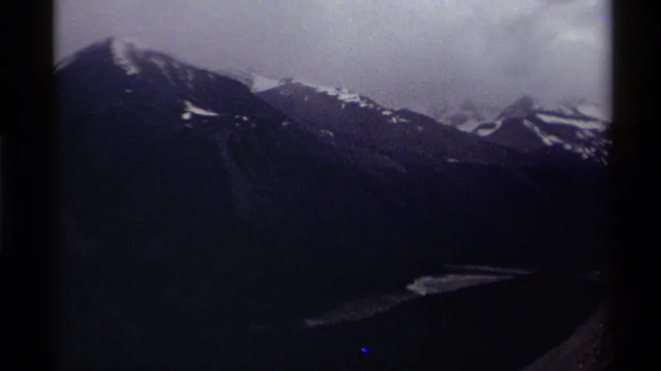 1979 Canada 8mm frame with clouded peaks, dark evergreen slopes, and a pale water or ice strip near the valley floor.