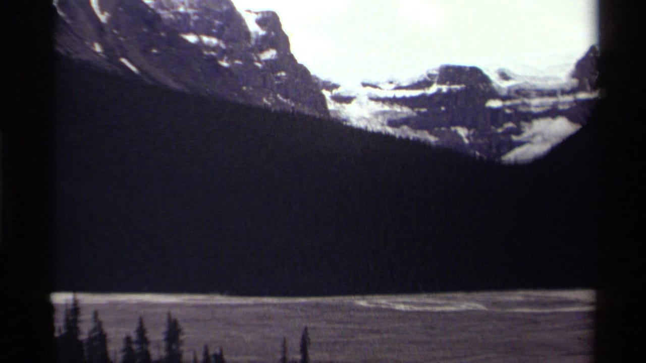 1979 8mm home-movie frame from Canada showing snow-streaked mountains, dark evergreen forest, and a lake under bright cloud cover.