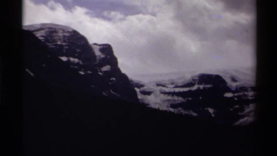 Soft 8mm view of a Canadian mountain slope with snow patches, silhouetted forest below, and cloud cover across the sky.