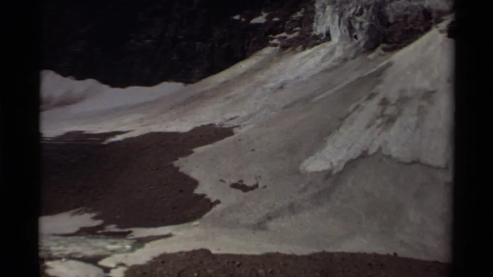 1979 Canada 8mm frame showing a steep snow-streaked slope, exposed rock, gravel, and dark meltwater below.