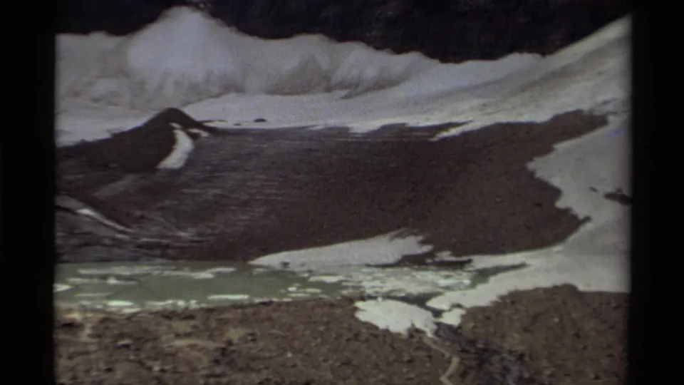 1979 Canada 8mm frame showing a cloudy mountain landscape with melting snow, dark rock, and a pale lake below.