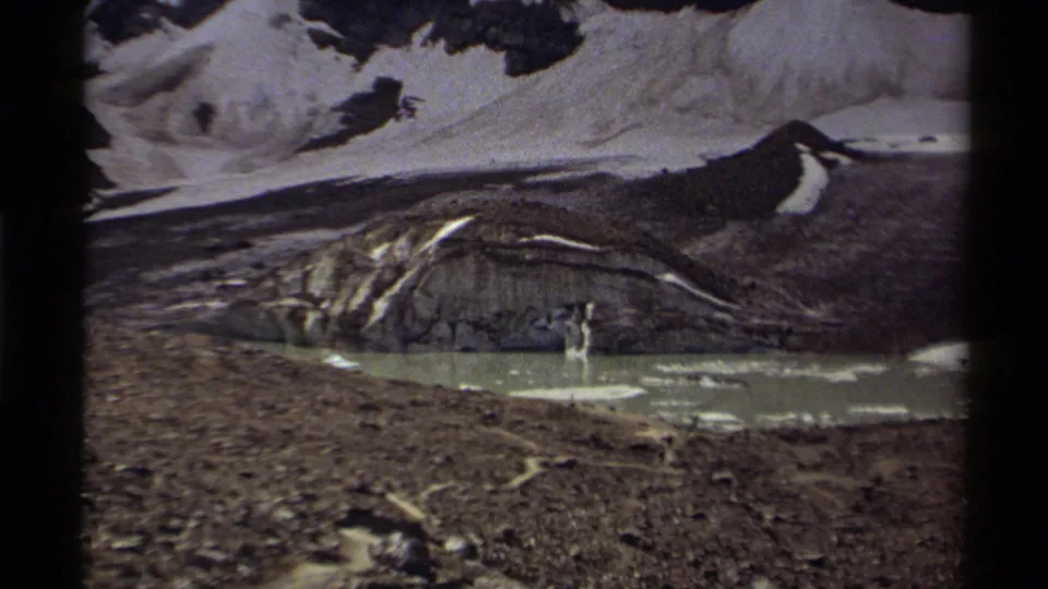 1979 Canadian 8mm frame showing exposed rock, snowfields, and a small pale meltwater pond at the base of a glacier-like slope.