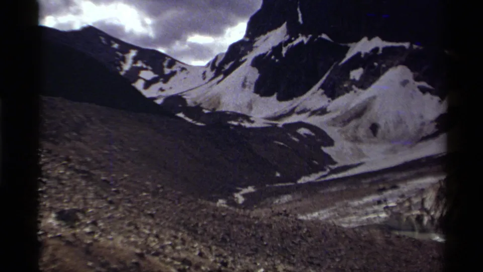 1979 Canadian 8mm frame looking across a rugged mountain valley with snow patches, dark rock faces, cloudy sky, and rough terrain.
