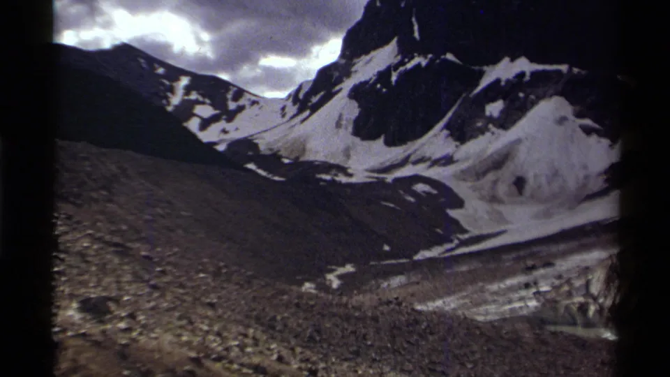 Canadian 1979 home-movie frame showing snow-covered peaks, exposed dark rock, and a glacier-like basin under a cloudy sky.