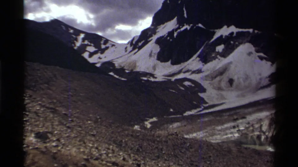 1979 Canadian home-movie frame showing snow-streaked mountain peaks, dark slopes, and a cloudy sky over a rocky valley.