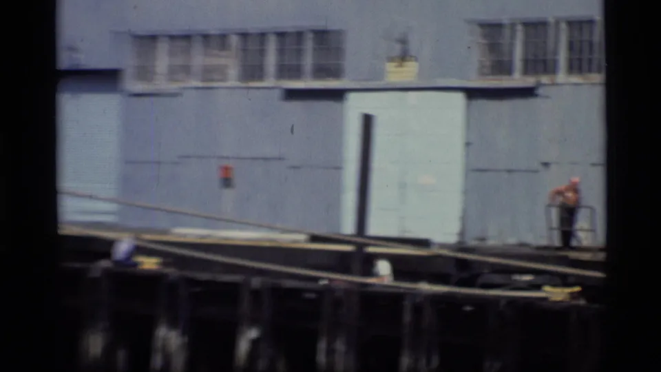 1977 Alaska home-movie frame showing a worker near gray waterfront buildings and pier pilings at a dock.
