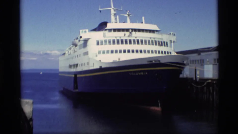 1977 Alaska frame showing the Columbia docked at a wooden pier, with open water and distant shoreline behind the vessel.