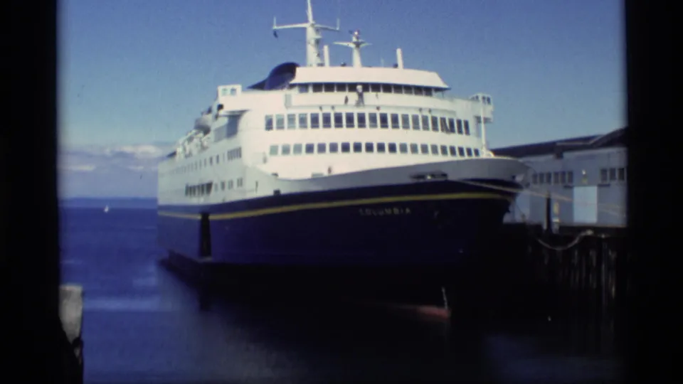 1977 Alaska home-movie frame showing the ferry Columbia docked at a pier on a calm bay under a bright sky.