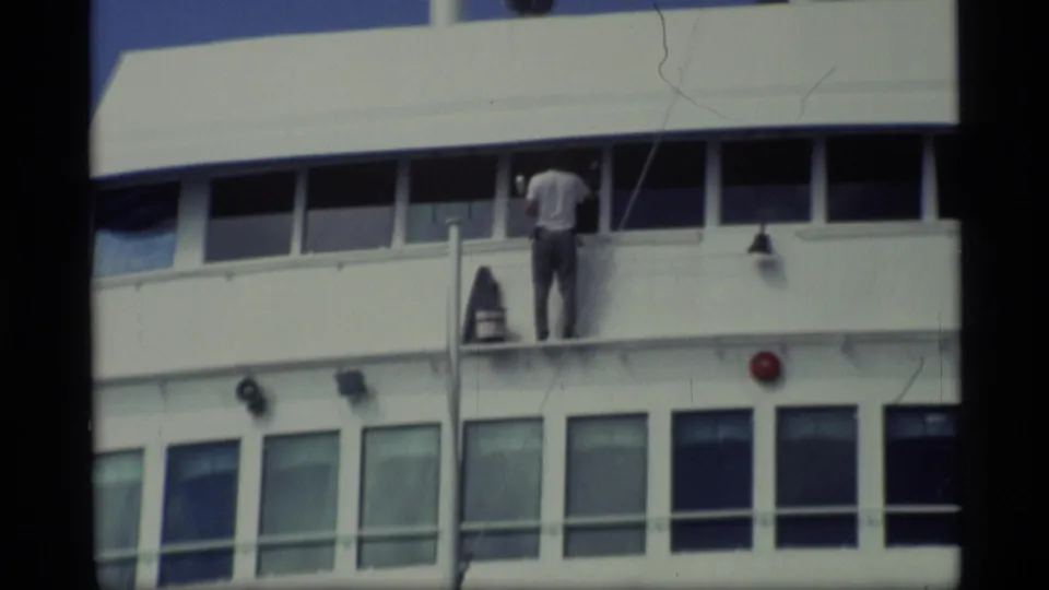 1977 Alaska home-movie frame with a crew member on the upper deck of the passenger ship near windows and railings.