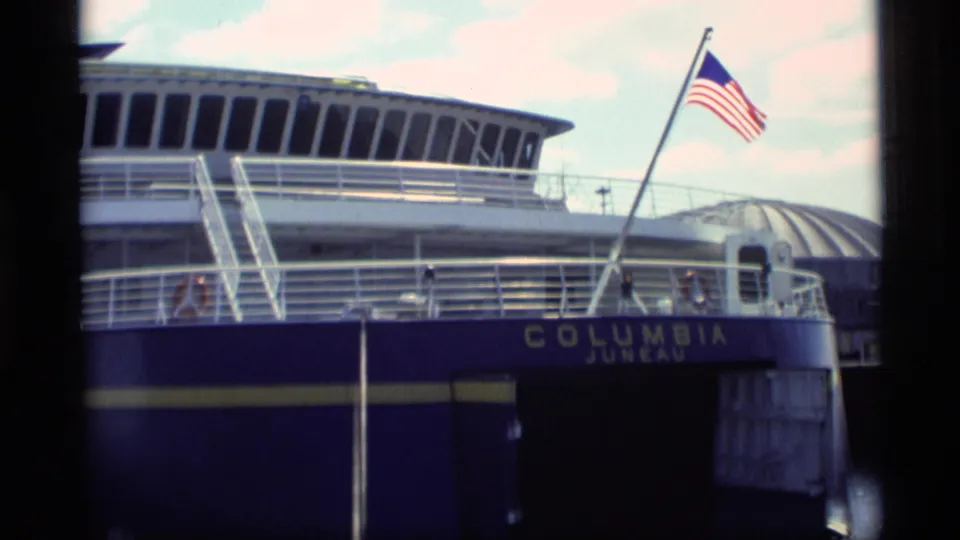 1977 Alaska ferry view with Columbia Juneau lettering, blue hull, railings, stairs, and an American flag flying from a mast.