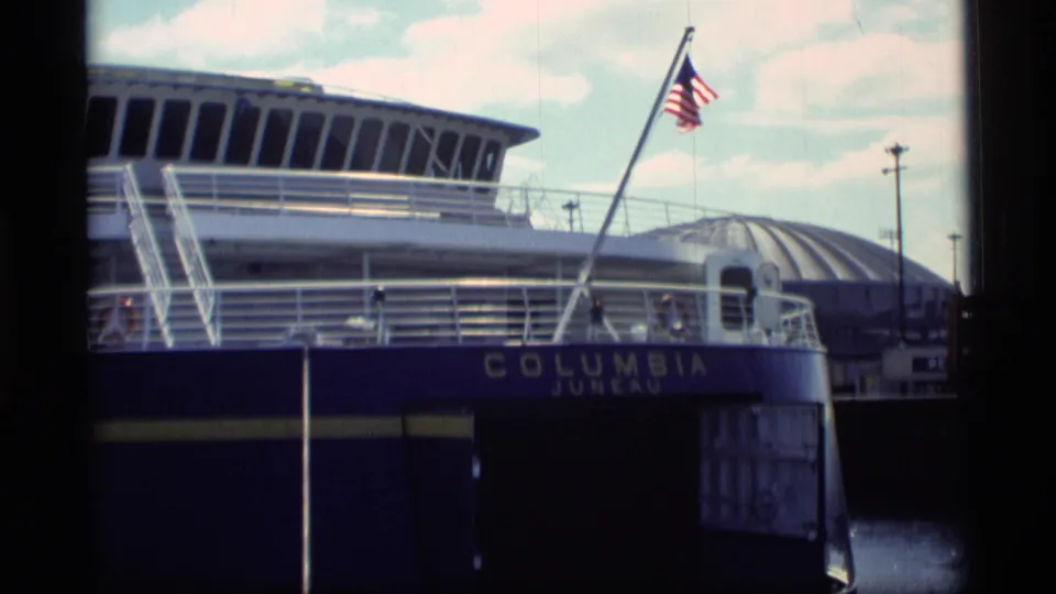 The Columbia Juneau ferry remains at dock, showing the flag, upper deck, railings, blue hull, and partial shoreline or port structure.