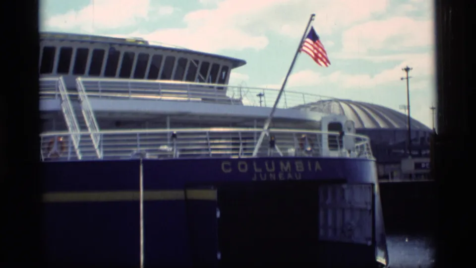 1977 8mm frame of the Columbia ferry docked near a pier, with an American flag moving in the wind above the visible ship name.
