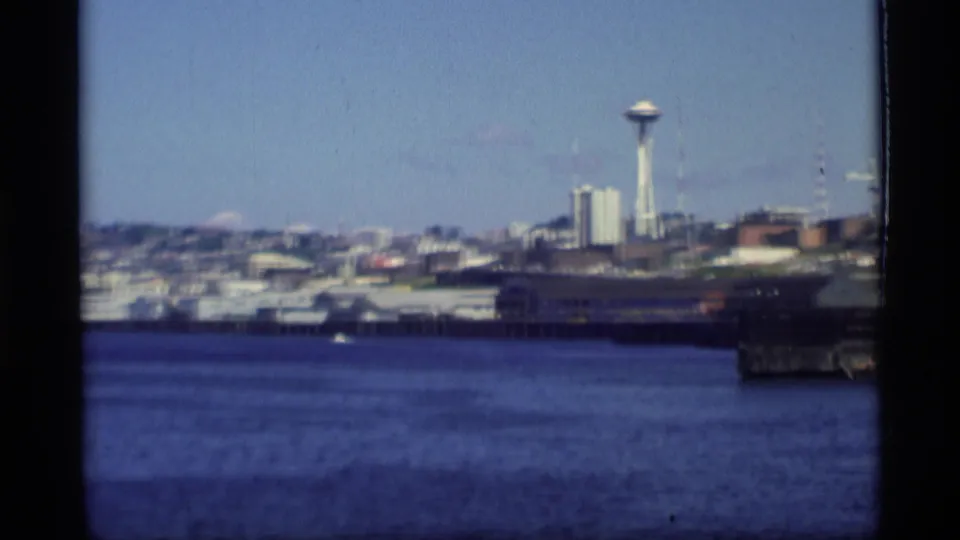 View across water toward the Seattle waterfront, with the Space Needle visible beyond shoreline buildings in soft 8mm focus.