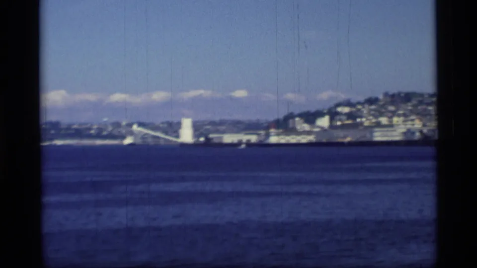1977 8mm Alaska view across blue ocean toward a distant hillside town, with a white shoreline tower visible near the water.