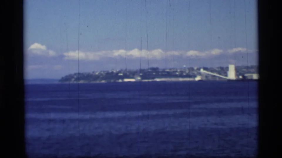 Metadata-supported Alaska coastal view from 1977, showing blue water, a distant hillside town, and a white tower near shore.
