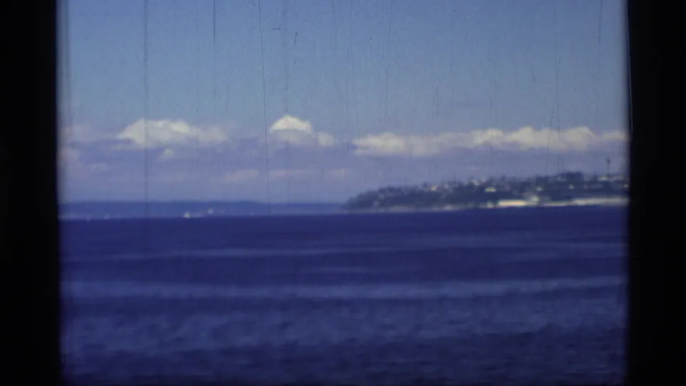 1977 8mm Alaska ocean view with a distant shoreline hillside at right, scattered clouds, and visible film wear.