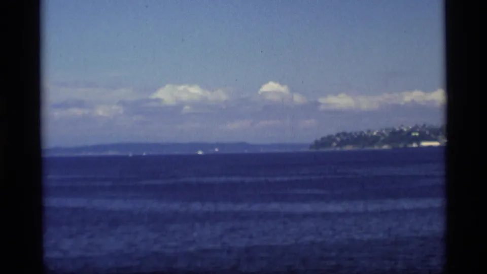 Vintage 8mm Alaska coastal view, with open blue water, distant landforms, and hillside buildings entering from the right.
