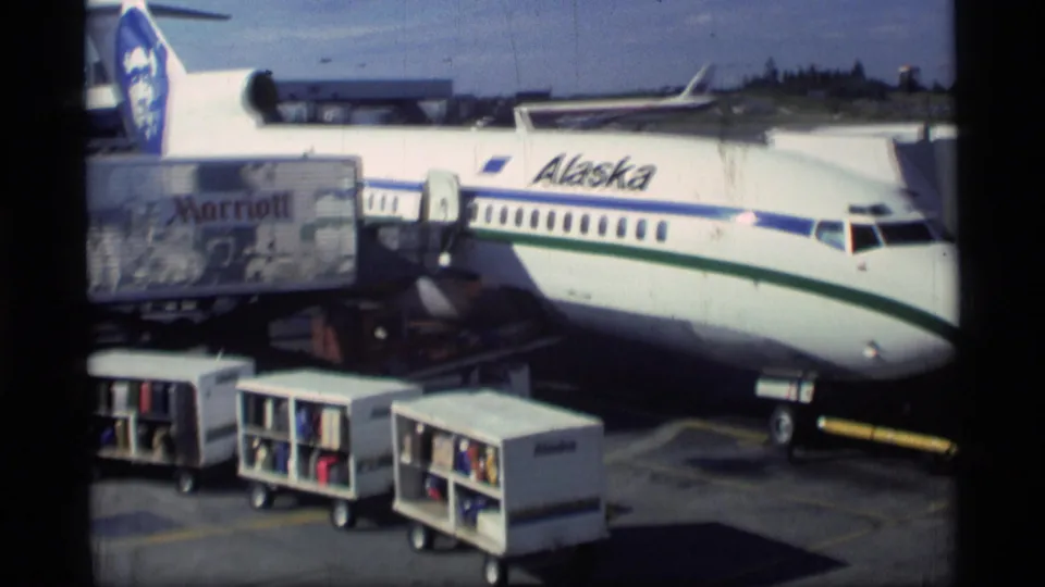 1977 Alaska airport ramp view showing an Alaska Airlines jet at a gate with service carts and a Marriott-branded container.