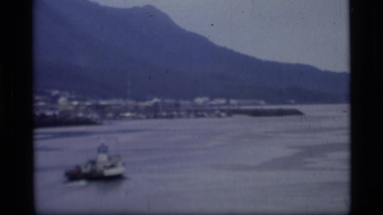 1977 Alaska home-movie view of a coastal harbor, mountains in the background, and a small boat moving across calm water.