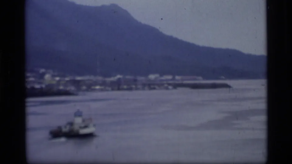 1977 Alaska home-movie view of a coastal harbor, mountains in the background, and a small boat moving across calm water.
