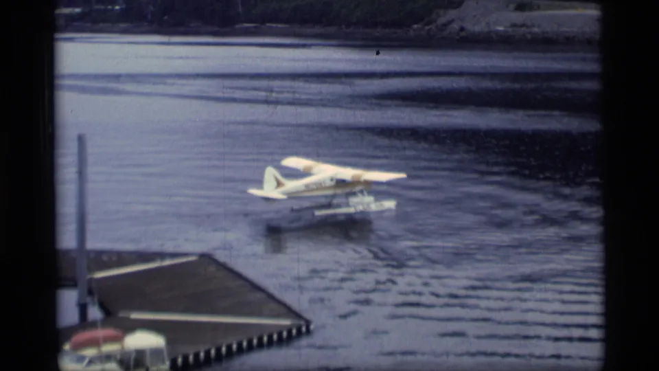 Floatplane taxis across calm harbor water near a wooden dock and red-roofed waterfront shed in 1977 Alaska footage.