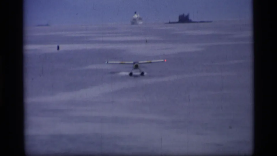 Floatplane lifts from the harbor surface in Alaska, leaving a faint wake with distant boats or ship silhouettes on the horizon.