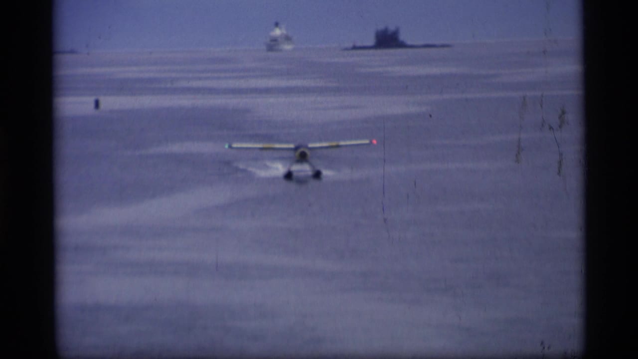 1977 Alaska 8mm frame showing a small floatplane accelerating on calm water, with a ship and hazy shoreline in the distance.