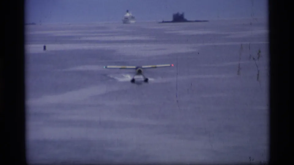 1977 Alaska 8mm frame showing a small floatplane accelerating on calm water, with a ship and hazy shoreline in the distance.