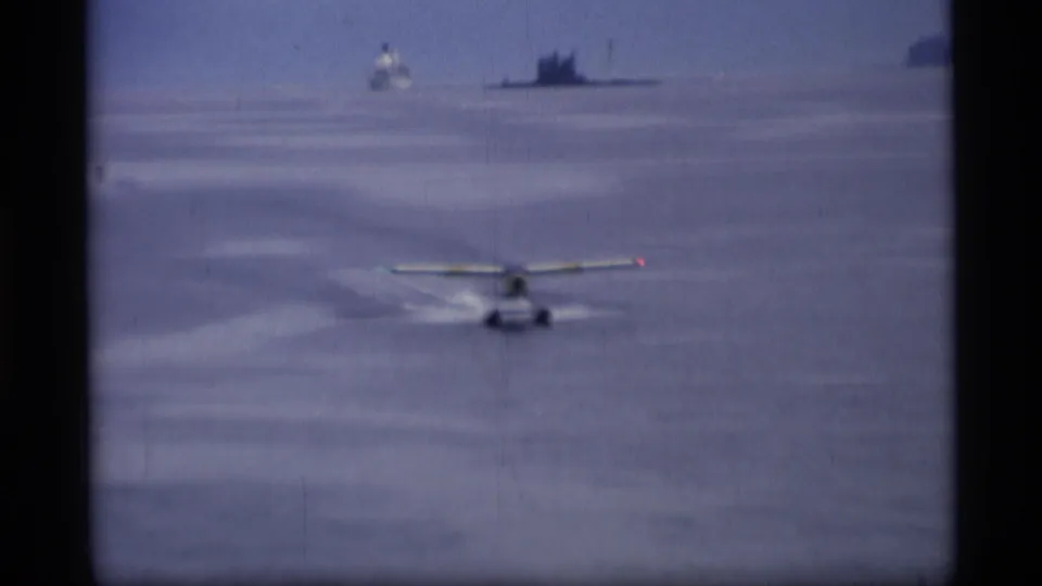 Small seaplane seen from behind during its water run in Alaska, with open water, distant vessel, and muted 8mm color.