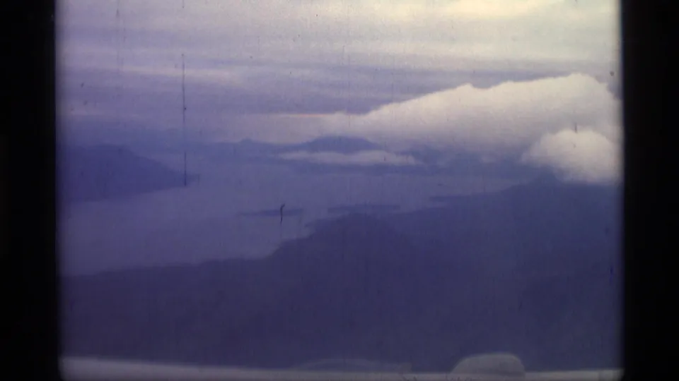 Final 1977 Alaska 8mm frame of a hazy coastal mountain view under bright low clouds and muted blue-purple film tones.
