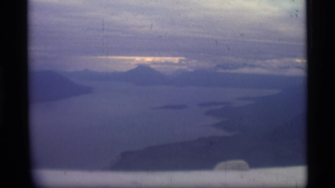 Soft 1977 8mm Alaska travel frame showing a hazy mountain and water view under clouded sky.
