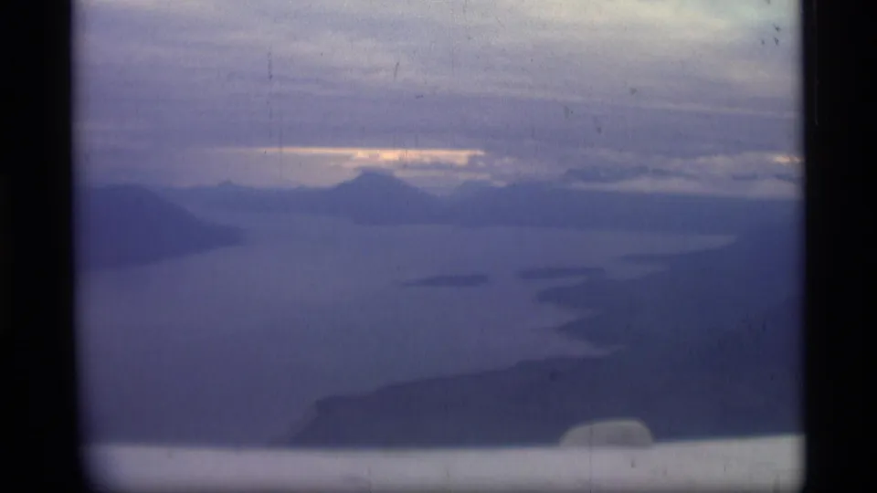 Soft 1977 8mm Alaska travel frame showing a hazy mountain and water view under clouded sky.