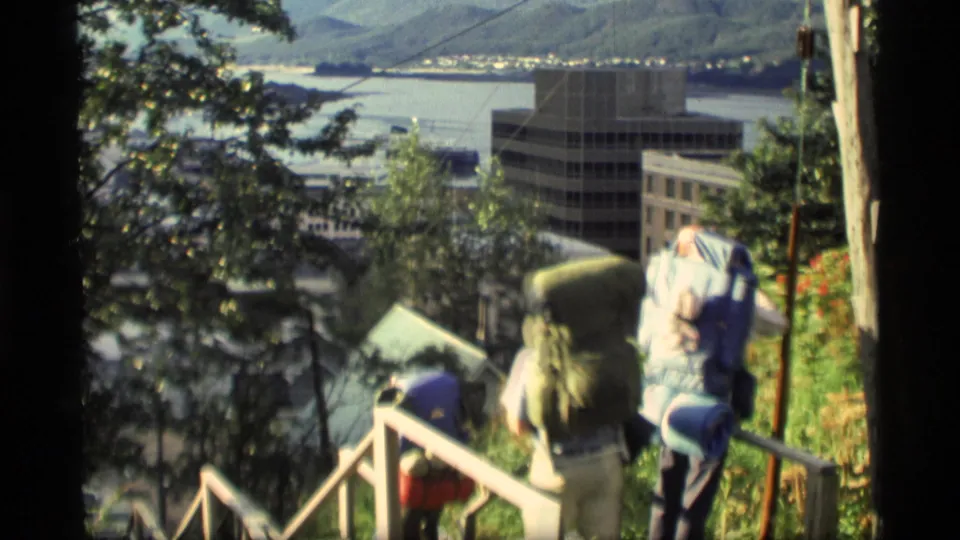 1977 Alaska home-movie frame of backpackers on a wooden stairway overlooking buildings, water, and mountains.