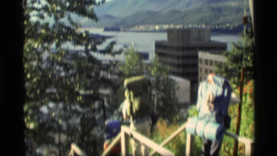 1977 Alaska 8mm frame showing travelers with camping packs descending stairs toward a waterfront city below.