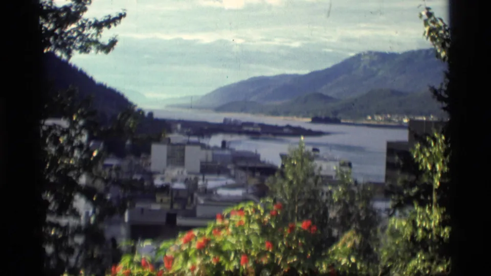 1977 Alaska home-movie frame looking over a waterfront city, bay, mountains, trees, and red flowers.