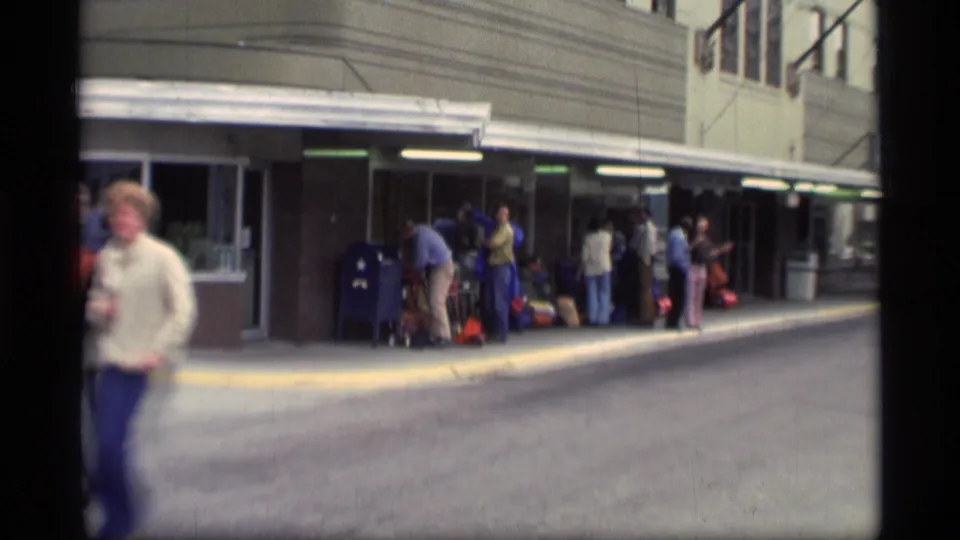 1977 Alaska street scene with adults, luggage, a covered sidewalk, and a passerby in soft focus.