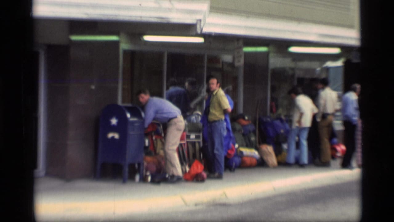 Travelers stand outside a terminal with luggage under a covered curbside area.