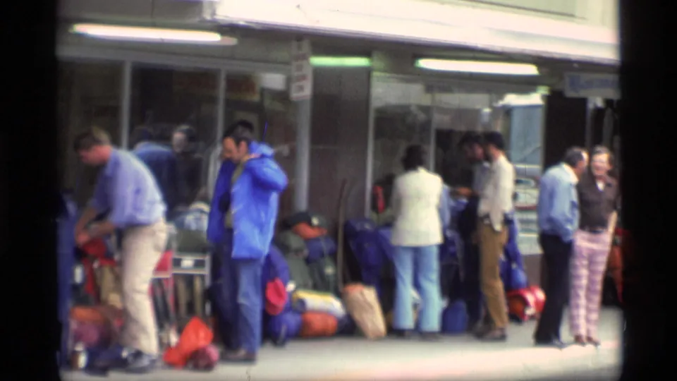 Curbside terminal scene in Alaska, 1977, with travelers, luggage piles, a blue box, and canopy lighting.