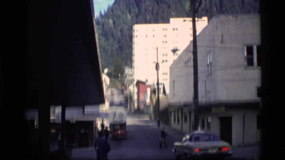 1977 Alaska street view with vintage cars, pedestrians, low buildings, and forested mountains in the background.