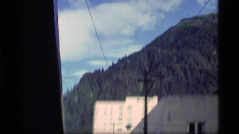 Mountain-backed Alaska view from 1977 home-movie footage, with a pale building in the foreground and overhead utility line.