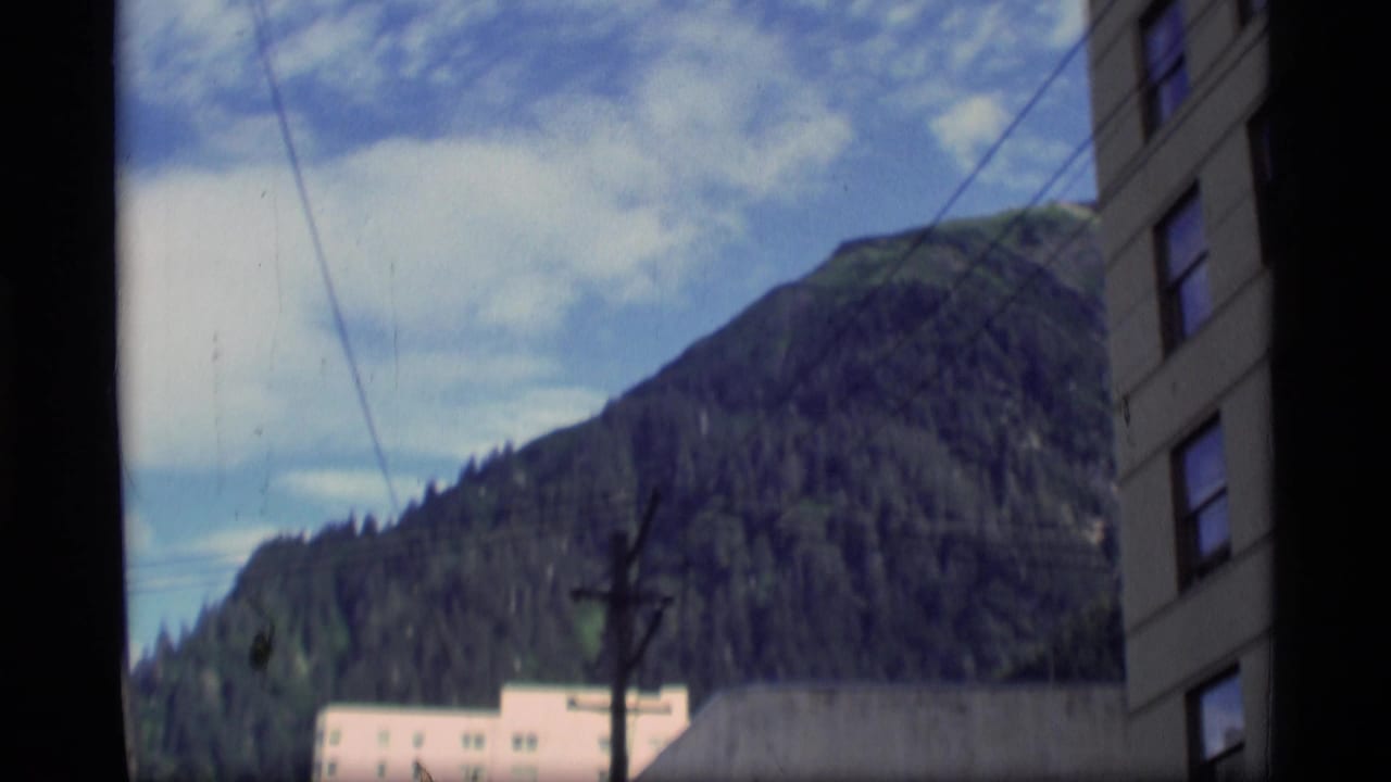 1977 Alaska home-movie frame showing a wooded mountain rising behind town buildings and utility lines.