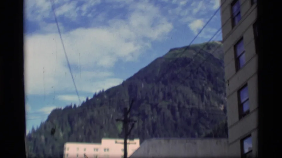 1977 Alaska home-movie frame showing a wooded mountain rising behind town buildings and utility lines.