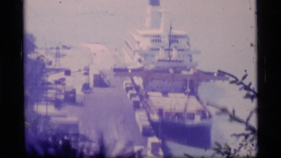 1977 Alaska harbor view with a large docked vessel, pier equipment, water, and soft overexposed home-movie color.