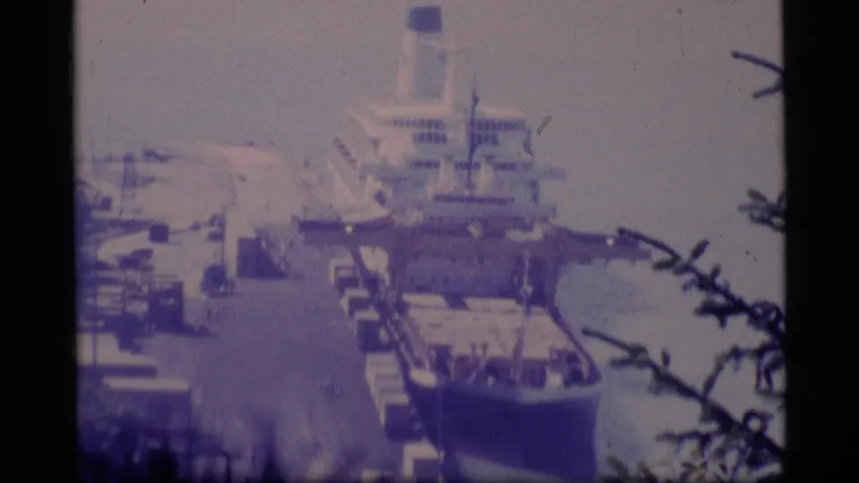 1977 Alaska harbor frame showing a large vessel moored beside a pier, with water and dark branches in the foreground.