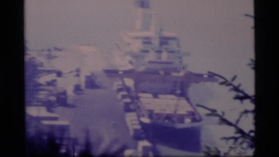 1977 Alaska home-movie frame showing a large moored vessel and dock area viewed from above through trees.
