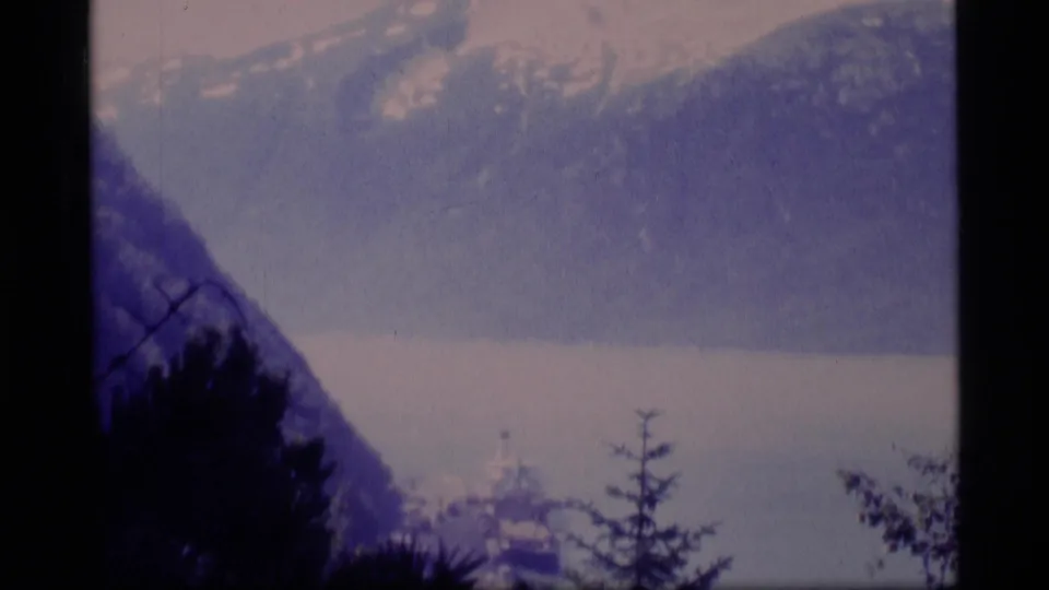 1977 Alaska travel footage frames a ship on the water against mountain ranges, with evergreen branches crossing the foreground.