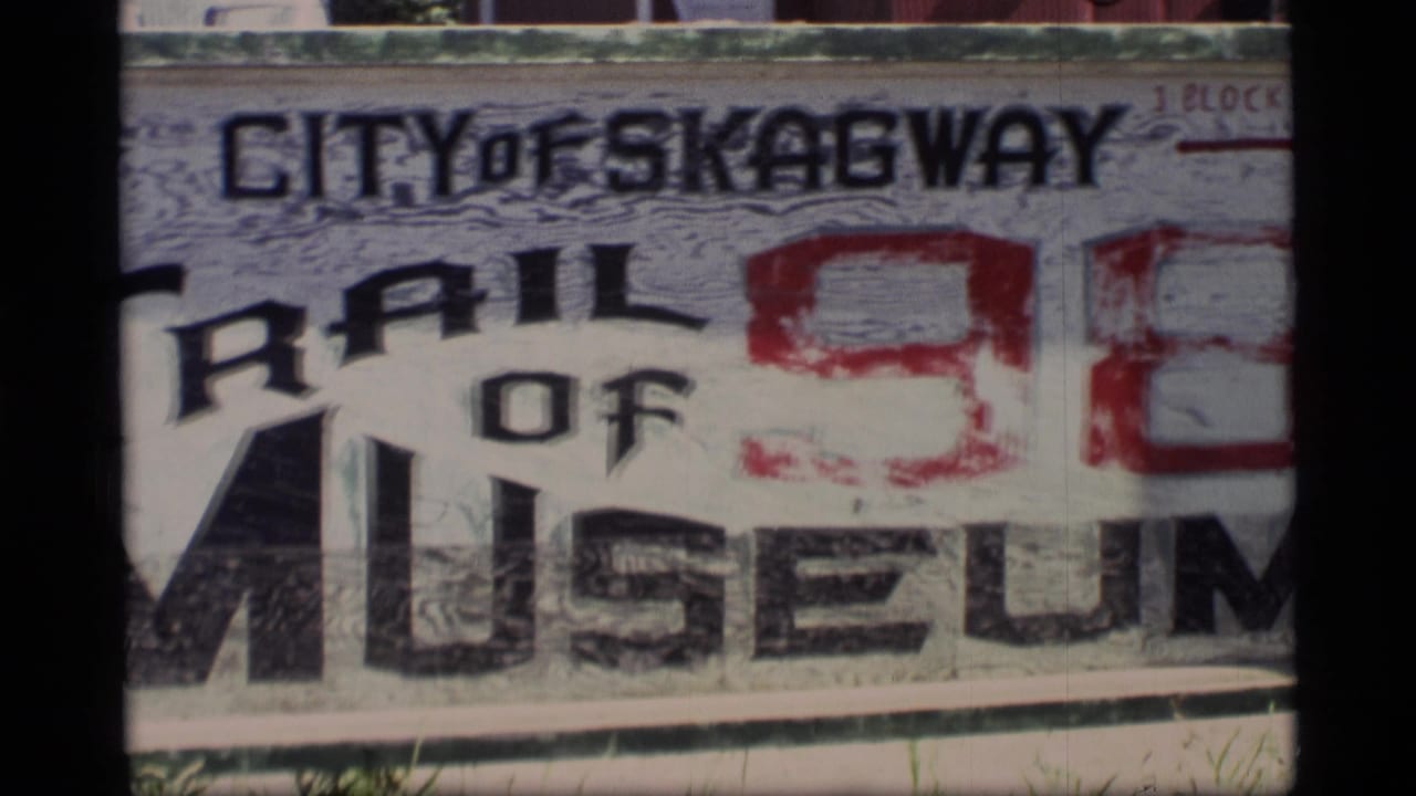 1977 Alaska home-movie frame showing a weathered Trail of 98 Museum sign in Skagway with bold black and red lettering.
