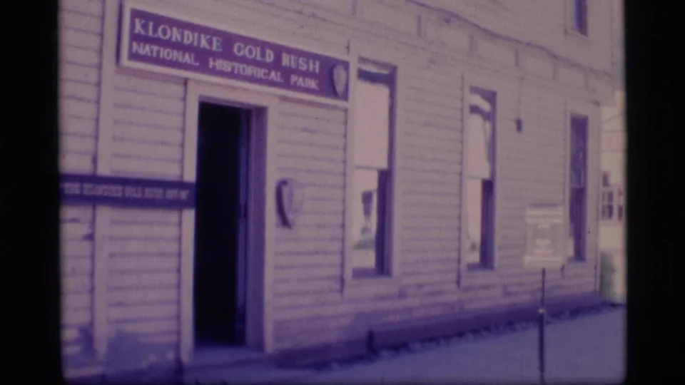 1977 Alaska frame showing a white clapboard building with a Klondike Gold Rush National Historical Park sign above an open doorway.