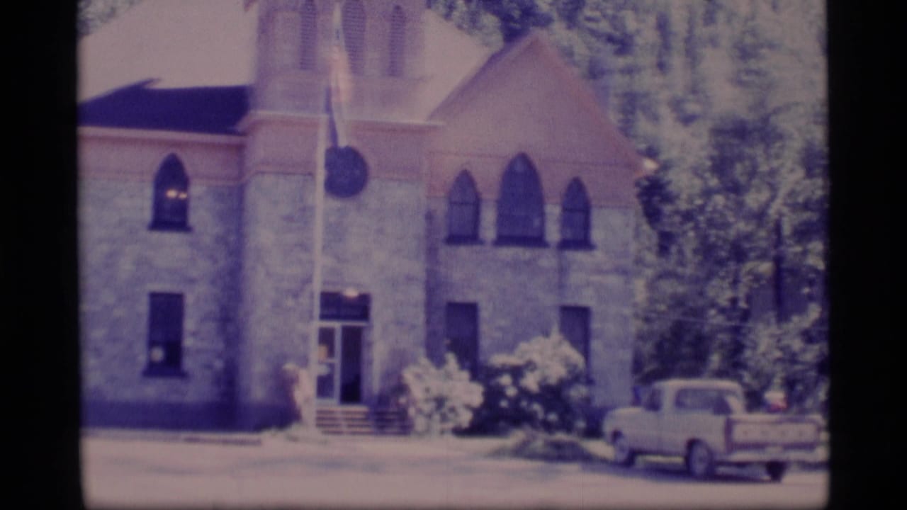 1977 Alaska 8mm frame showing a stone church-like building with Gothic-style windows, a flagpole, trees, and a light pickup truck.